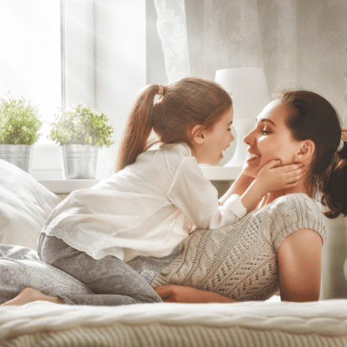 A mother is lying on a bed with her young daughter on top of her. The daughter is holding her mother's face with her hands as they both look at each other and smile.
